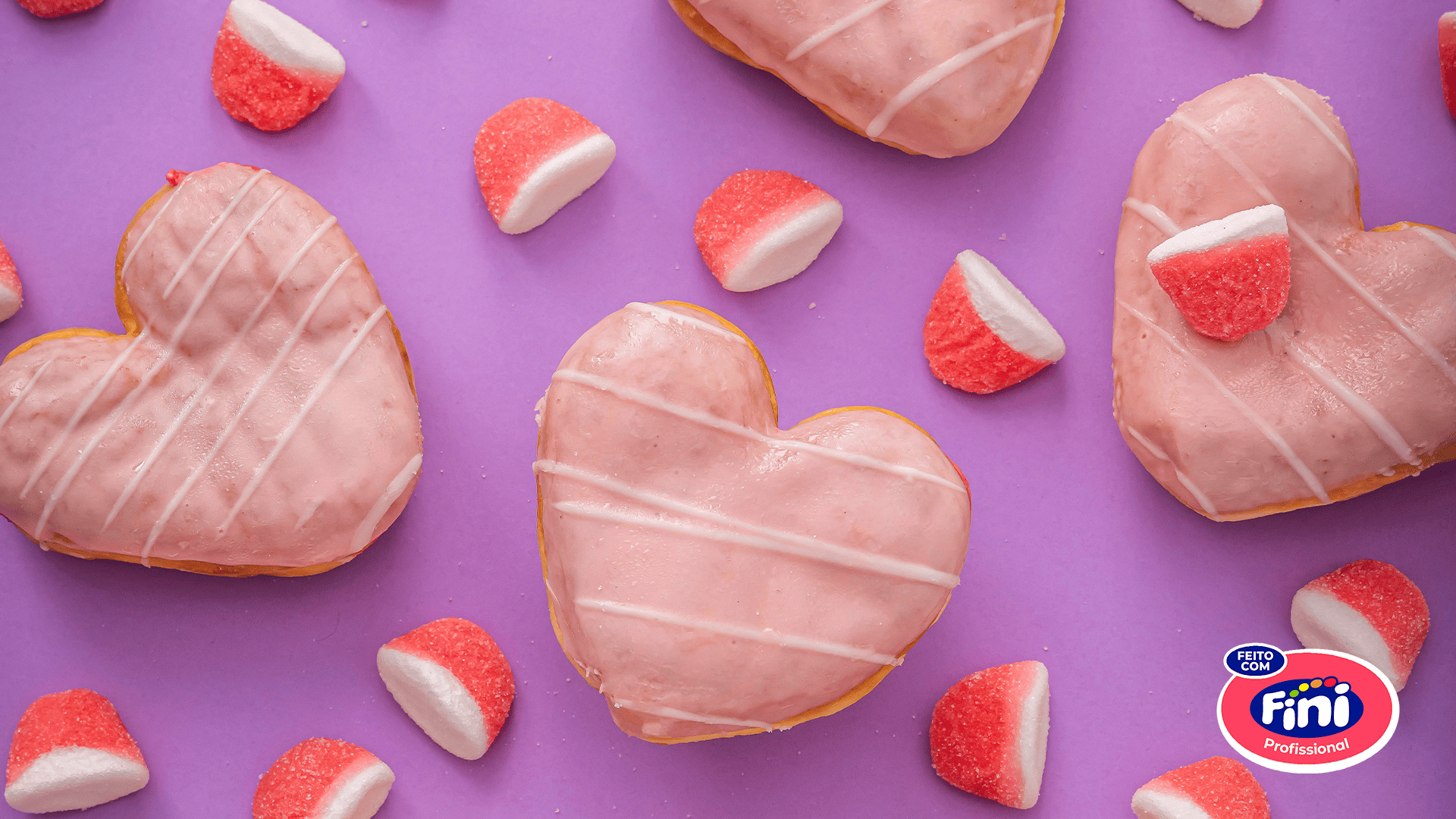 Donuts de coração rosa com glacê branco e balas Fini coloridas espalhadas sobre fundo roxo, destacando o lançamento irresistível da padaria Pão de Açúcar.