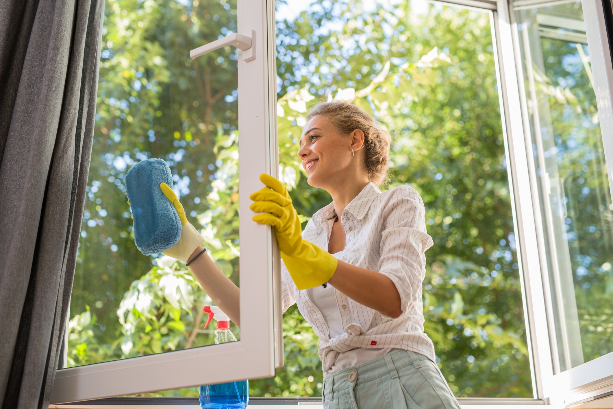 Mulher limpando janela com luvas amarelas e esponja azul durante a faxina de primavera em casa, preparando o ambiente para a mudança de estação