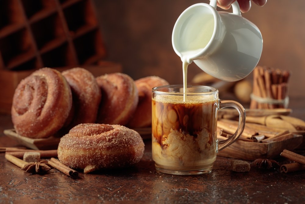 Jarro despejando leite quente em cappuccino cremoso ao lado de donuts açucarados, canela e grãos de café