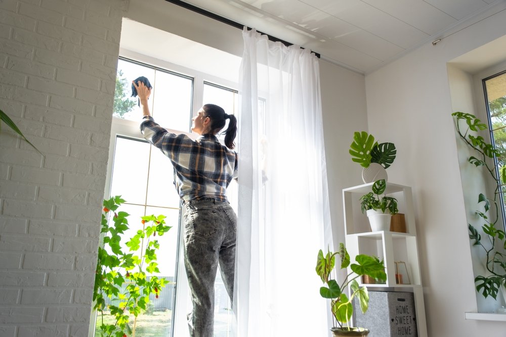 Mulher com camisa xadrez limpando janela de casa enquanto organiza plantas verdes, demonstrando cuidados com a casa na mudança de estação