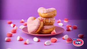 Pilha de donuts de coração rosa com glacê em um prato rosa, cercados por balas Fini, destacando a irresistível novidade da padaria Pão de Açúcar.
