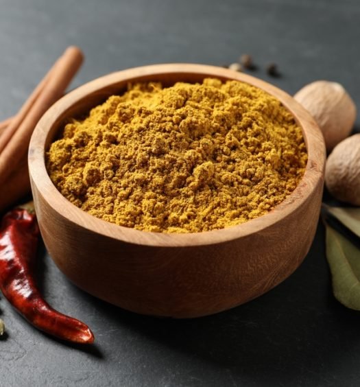 Pó de curry amarelo vibrante em uma tigela de madeira com especiarias secas ao redor, destacando a cúrcuma.