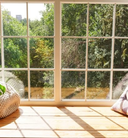 Sala iluminada pela luz natural com grande janela de vidro, almofadas em tons neutros e plantas verdes, representando ambiente aconchegante para preparação da mudança de estação