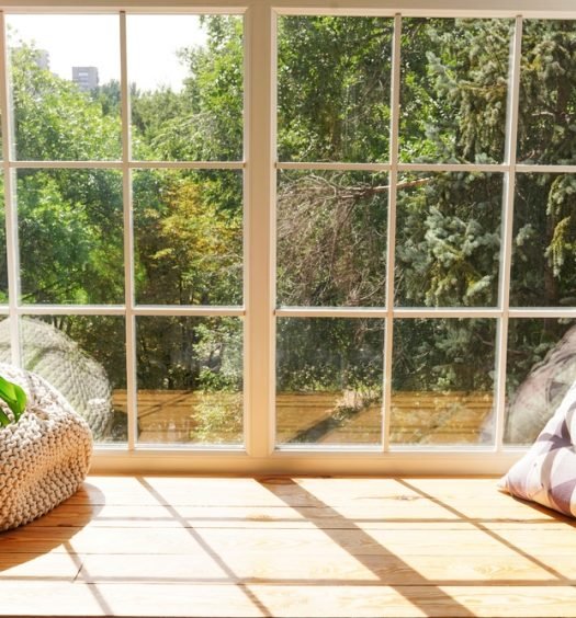 Sala iluminada pela luz natural com grande janela de vidro, almofadas em tons neutros e plantas verdes, representando ambiente aconchegante para preparação da mudança de estação