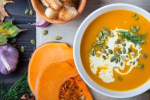 Tigela de sopa cremosa de abóbora com caldo branco e sementes de abóbora decorando o topo, acompanhada de abóbora fresca cortada e pão torrado