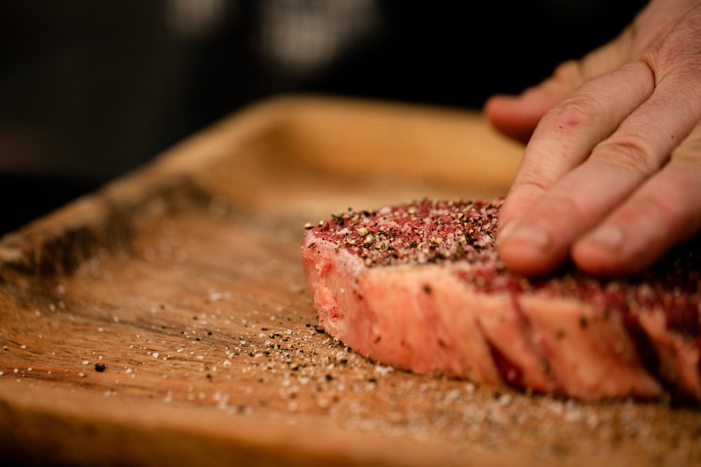 Close-up de mão aplicando tempero em pó e especiarias sobre fatia de carne em tábua de madeira, mostrando técnica de preparo detalhada para receitas saborosas