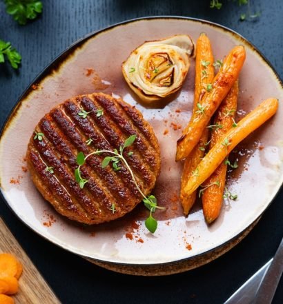Hambúrguer vegetariano caseiro grelhado servido com cenouras assadas e flor de cebola em prato rústico sobre mesa escura