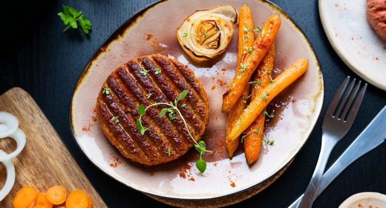 Hambúrguer vegetariano caseiro grelhado servido com cenouras assadas e flor de cebola em prato rústico sobre mesa escura