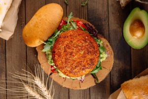 Hambúrguer vegetariano de grão-de-bico caseiro servido em pão com rúcula, guacamole e pimenta vermelha sobre tábua de madeira rústica com abacate ao lado
