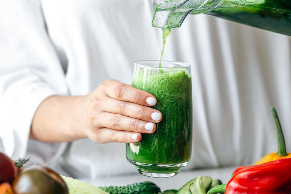 Pessoa servindo suco verde detox em copo de vidro cercado por legumes e verduras coloridos simbolizando bebida queridinha da saúde