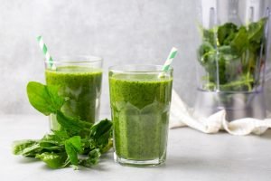 Suco verde detox em dois copos de vidro com folhas de couve frescas ao lado e liquidificador ao fundo mostrando bebida saudável