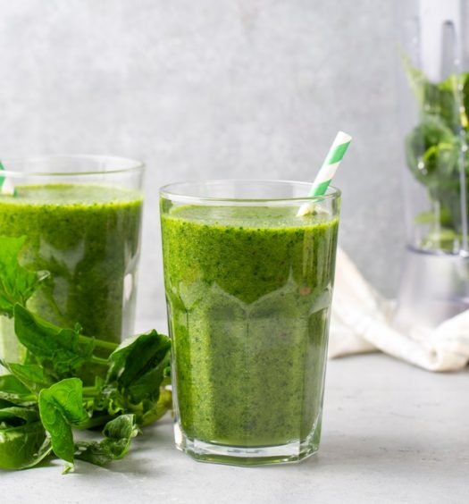 Suco verde detox em dois copos de vidro com folhas de couve frescas ao lado e liquidificador ao fundo mostrando bebida saudável