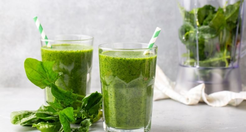 Suco verde detox em dois copos de vidro com folhas de couve frescas ao lado e liquidificador ao fundo mostrando bebida saudável