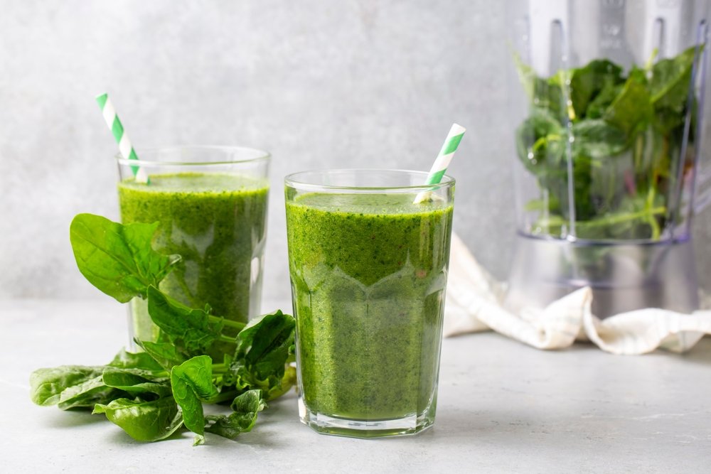 Suco verde detox em dois copos de vidro com folhas de couve frescas ao lado e liquidificador ao fundo mostrando bebida saudável