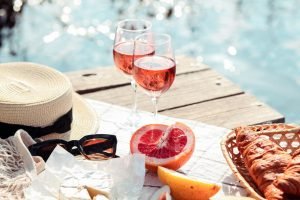 Piquenique de verão com duas taças de vinho rosé frutado à beira da água, chapéu de palha, óculos de sol e frutas cítricas.