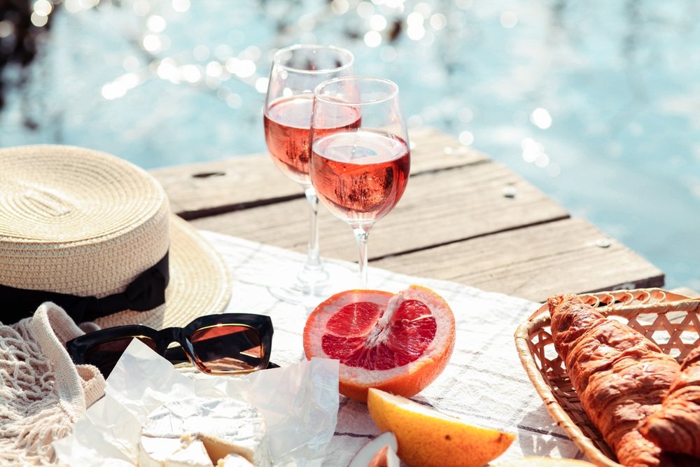 Piquenique de verão com duas taças de vinho rosé frutado à beira da água, chapéu de palha, óculos de sol e frutas cítricas.