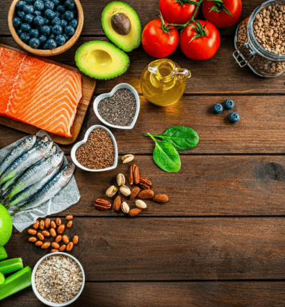 Vista superior de uma mesa de madeira com salmão, sardinhas, abacate, nozes, sementes de chia, linhaça e vegetais diversos.