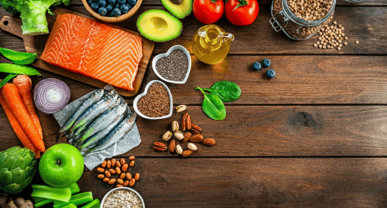 Vista superior de uma mesa de madeira com salmão, sardinhas, abacate, nozes, sementes de chia, linhaça e vegetais diversos.