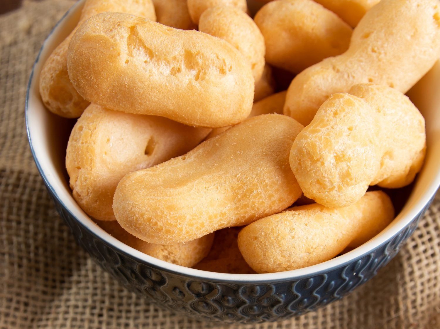 Close de um bowl de cerâmica azul com detalhes em relevo, cheio de biscoitos de polvilho doce em formato alongado e cilíndrico.
