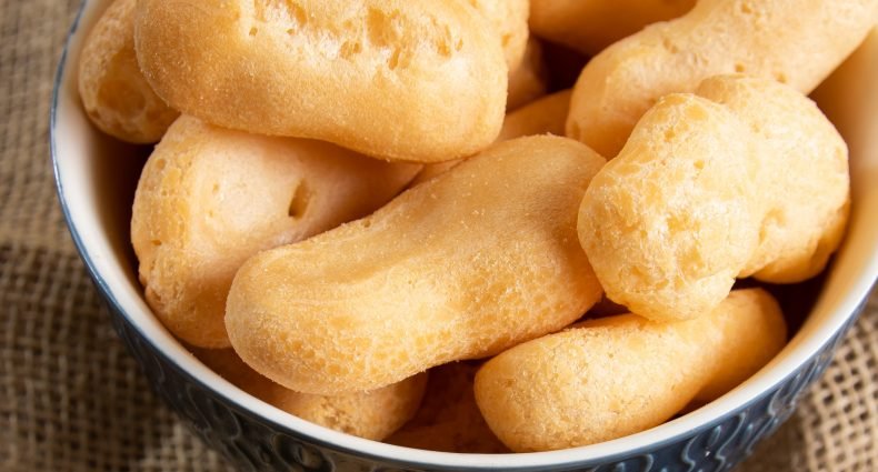 Close de um bowl de cerâmica azul com detalhes em relevo, cheio de biscoitos de polvilho doce em formato alongado e cilíndrico.
