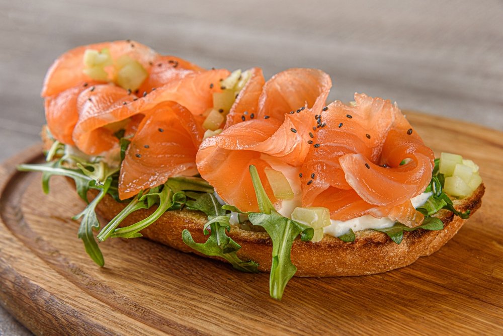 Sanduíche aberto com salmão defumado, rúcula e cubos de pepino sobre tábua de madeira, representando o caminho da rastreabilidade de pescados até a casa do consumidor.