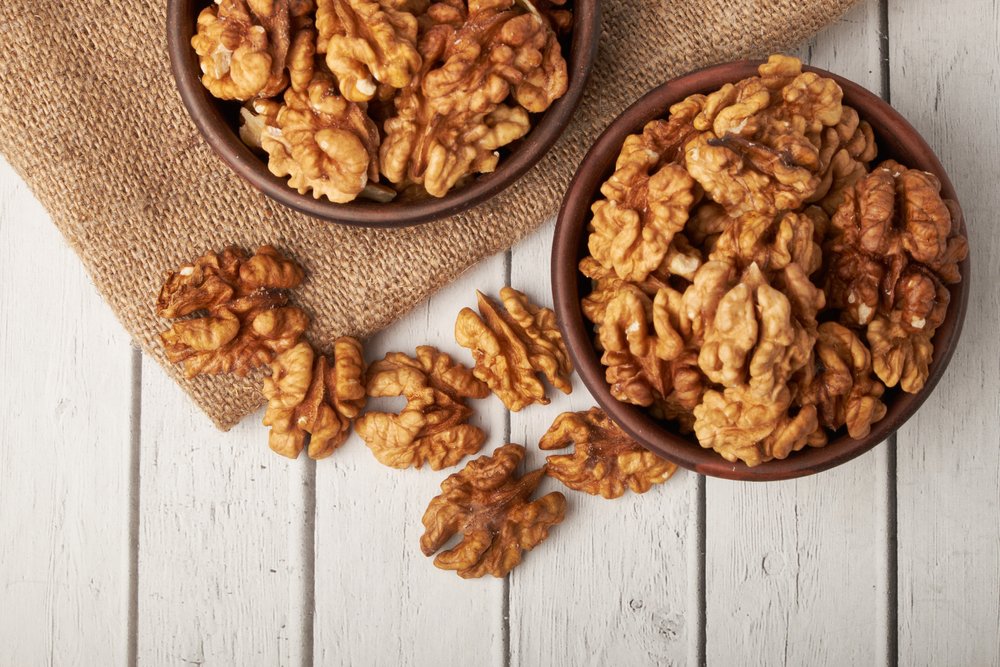 Dois recipientes de cerâmica marrom cheios de nozes sem casca sobre um fundo de madeira clara e tecido de juta.