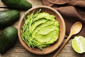 Tigela de madeira com purê de abacate (guacamole) decorado com brotos, cercada por abacates inteiros e meio limão.